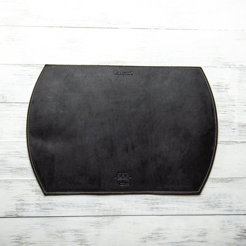 Straight edged oval personalized fine leather placemat in dark leather on a light wooden surface.