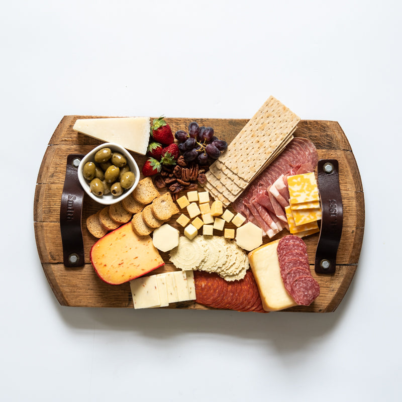 Serving tray made of Tennessee Whiskey Barrel wood and leather straps. Covered in a charcuterie board spread of meats and cheeses.