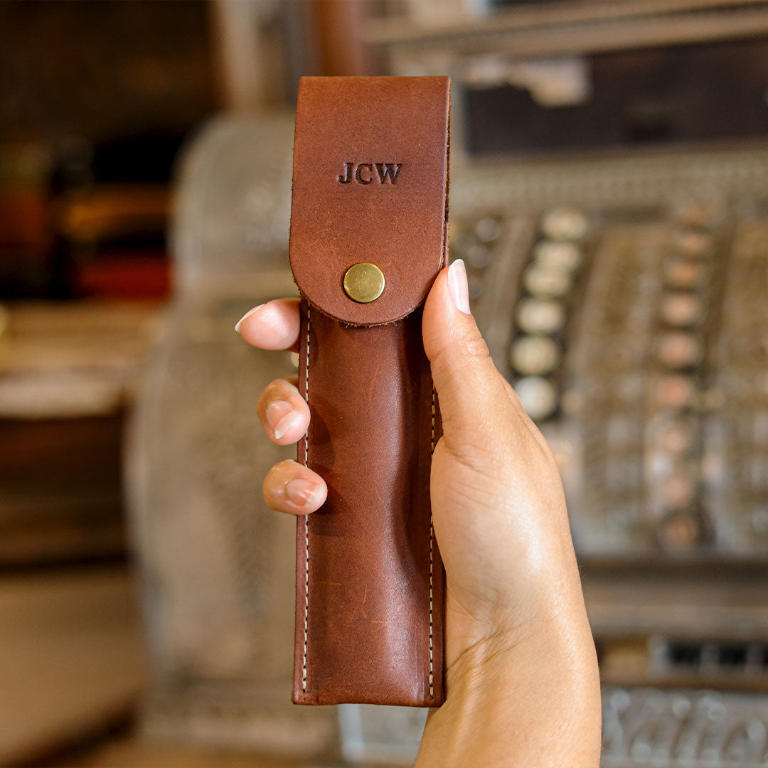 A hand holding a brown leather wallet or card holder with a snap tab and the initials 'JCW' embossed on the tab. The background is a blurred vintage-style interior with a cash register.