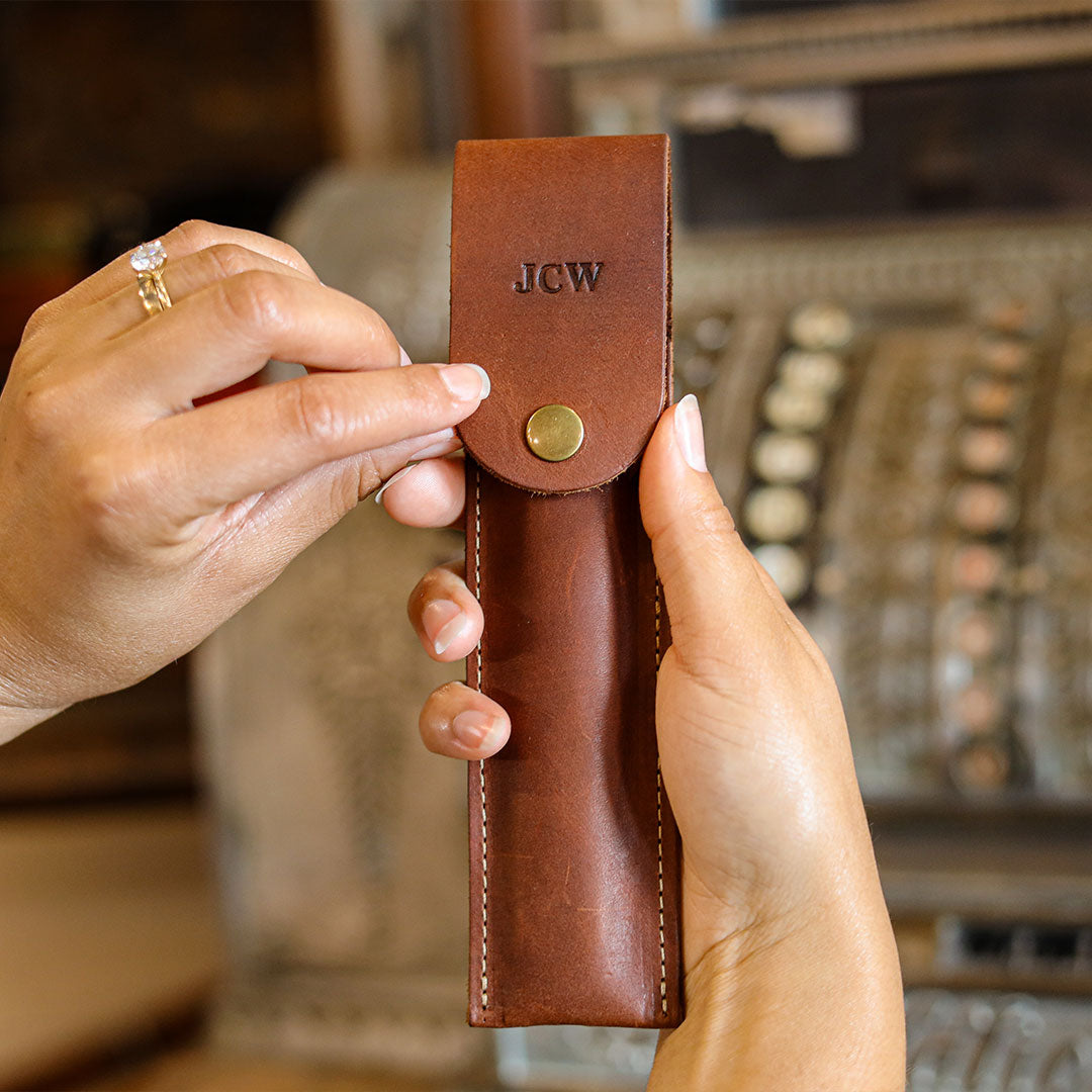 A person holds a brown leather snap-closure pouch with the initials 'J.C.