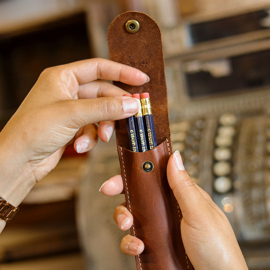 A close-up of a person holding a brown leather pencil case with two pencils inserted, set against a softly blurred background.